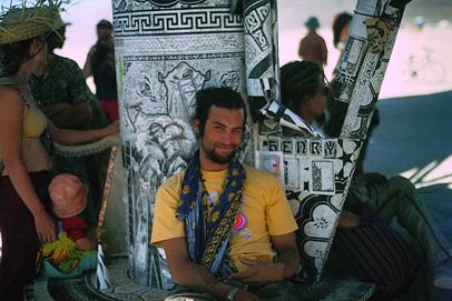 a smiling bearded man in a yellow shirt sits against black and white patterned panels, a woman and a baby beside him