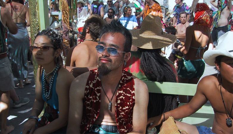 a man in blue sunglasses and a red patterned vest sits in a crowd in the sun, a woman in beads beside him