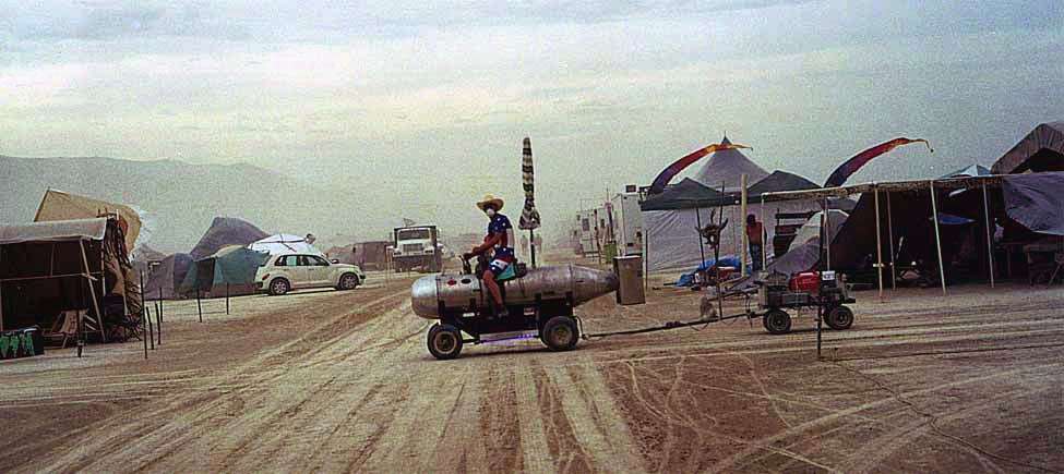 a man rides a small tank shaped vehicle across a dusty track between camps and tents