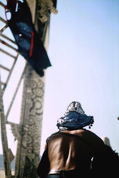 a person in a blue sequinned hat seen from behind, looking up at a patterned tower with a dark cloth hanging from it