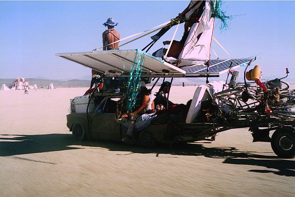 a car rebuilt as a winged machine with solar panels, people riding on it across the open desert floor