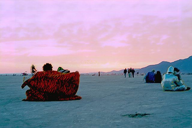 people wrapped in a tiger striped blanket and silver capes sit on the flat desert floor under a pink and violet sky