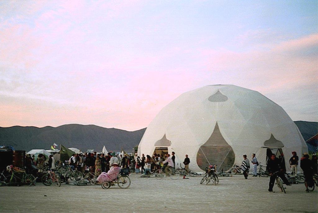 a white dome with pointed doorways at dusk, a crowd and bicycles around it under a pink sky