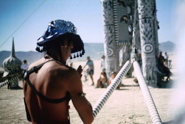 a person in a blue sequinned hat seen from behind, walking toward tall patterned pillars where people sit in the shade