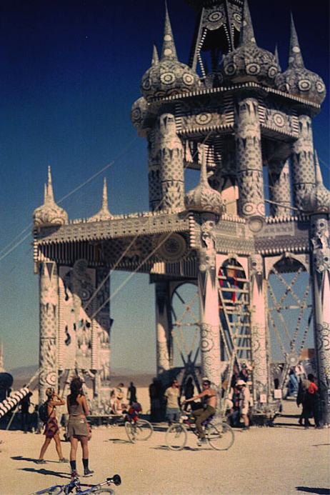 a tall tower of patterned spires and arches against a deep blue sky, people and bicycles at its foot