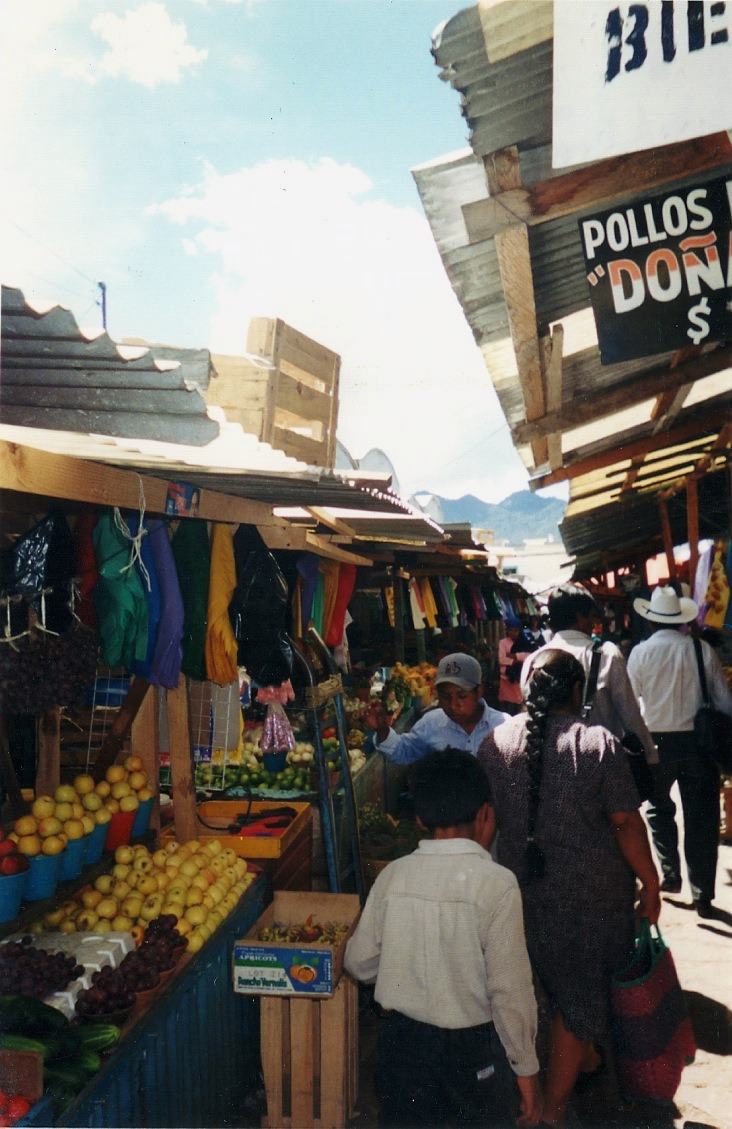 a market lane with fruit stalls and a POLLOS sign