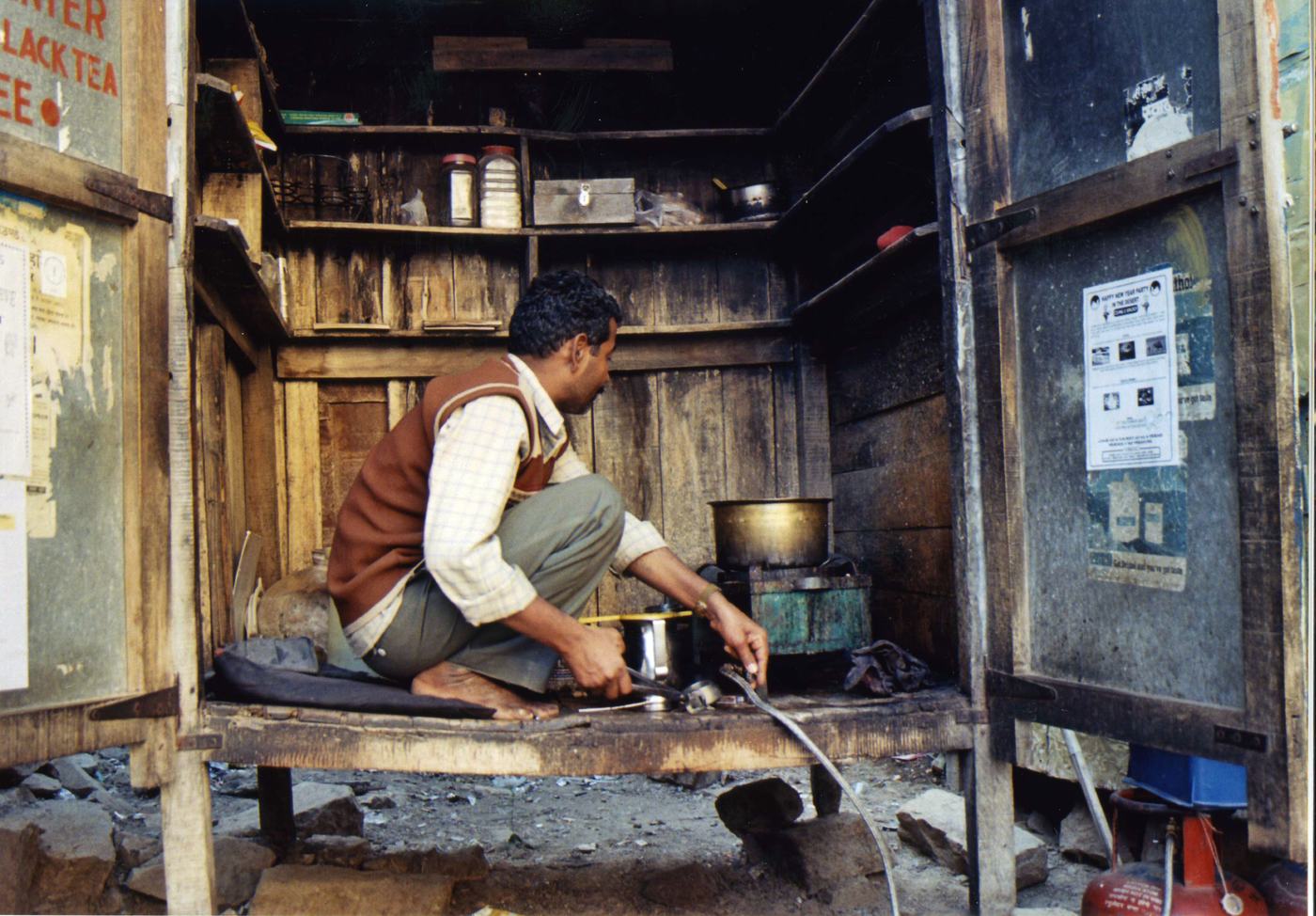 a man crouches on the floor of a wooden stall, making tea over a gas burner
