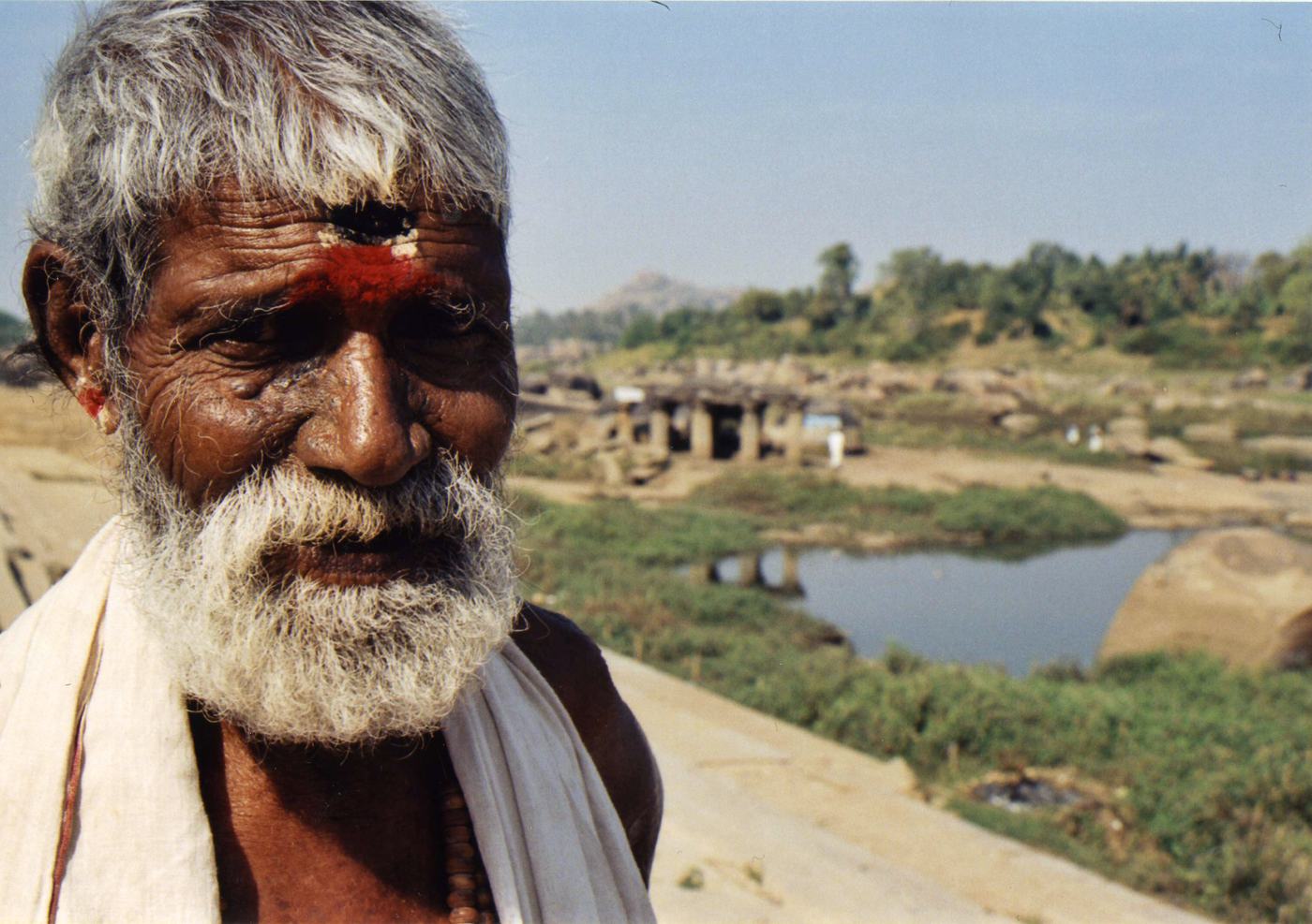an old man with a white beard and a red mark on his forehead, a river, boulders and stone ruins behind him at Hampi