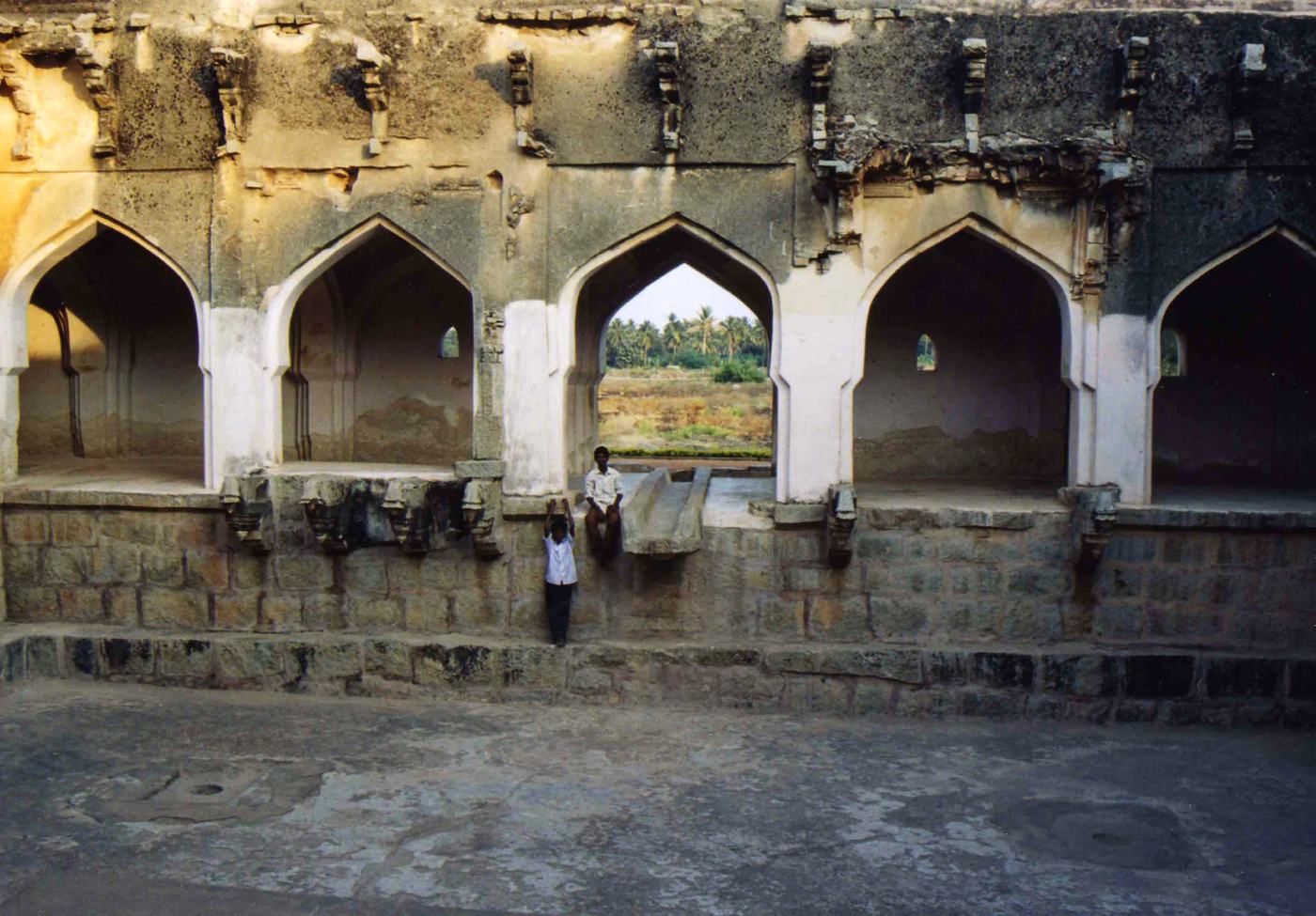 two boys stand below a row of pointed arches in an old stone building, palms seen through one arch