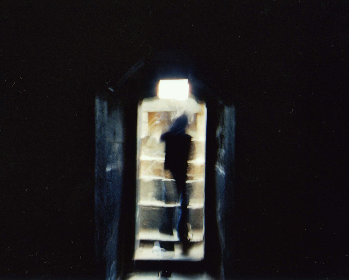 a blurred dark figure climbs a narrow flight of steps toward a bright opening, everything around it black