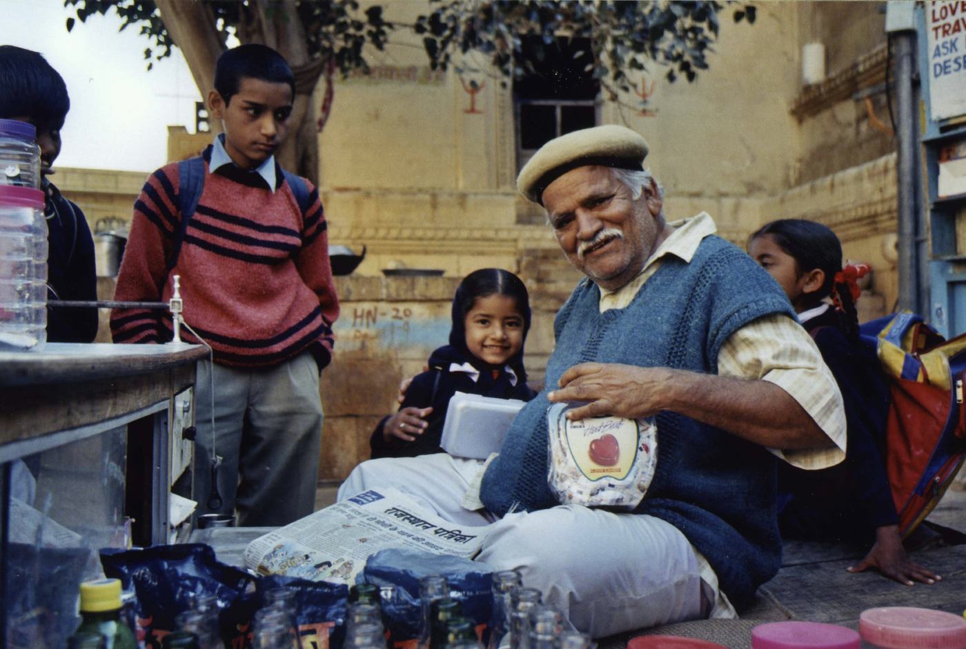 an old man with a white moustache and a flat cap sits cross legged at a street stand, smiling, schoolchildren around him
