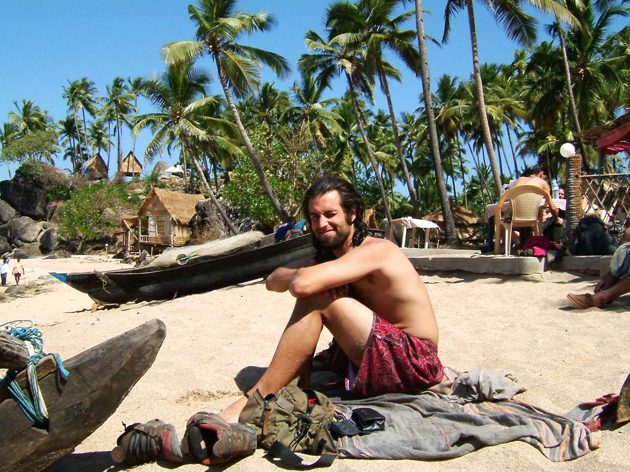 a smiling shirtless man with long hair sits on a blanket on the sand beside wooden boats, huts and coconut palms behind, in Goa