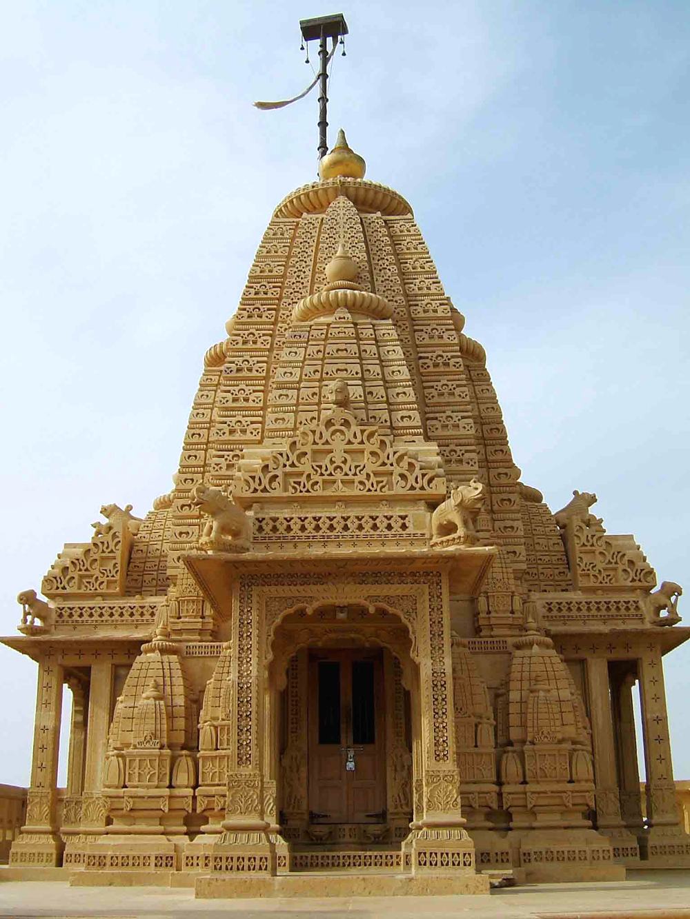 a small temple of carved golden sandstone with a tall spire, a flag on top against a pale sky