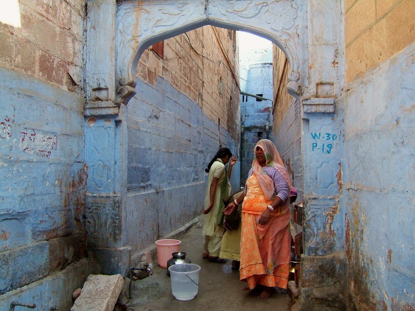 women in bright saris stand talking among buckets in a narrow blue painted alley under a carved stone arch