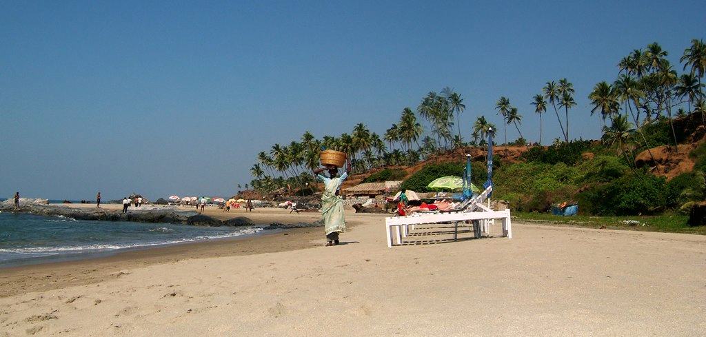 a woman in a green sari walks along a beach with a basket on her head, past white sunbeds, palms on the headland behind