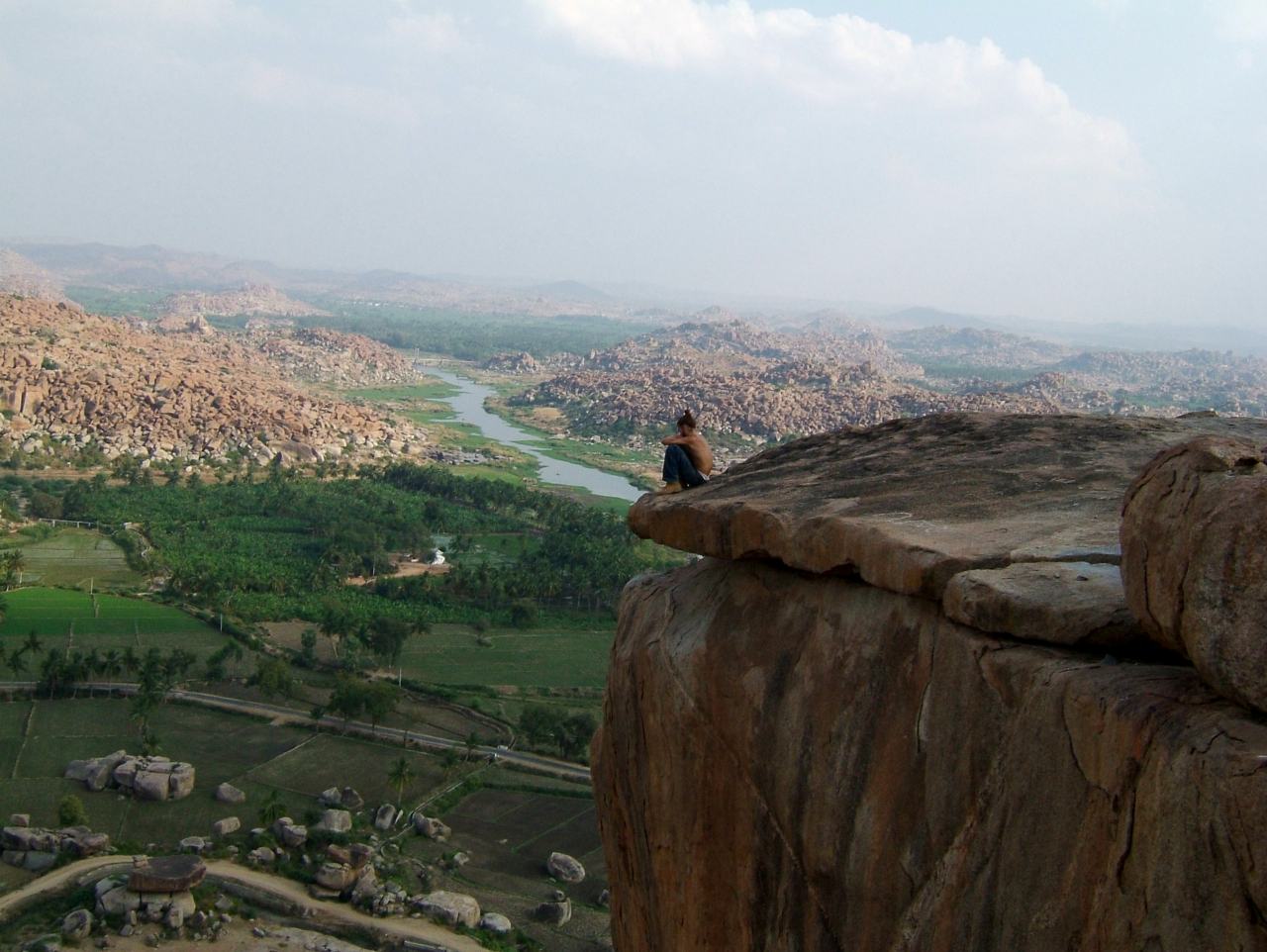 a person sits on the edge of a great boulder above a river valley of green fields and rock hills at Hampi