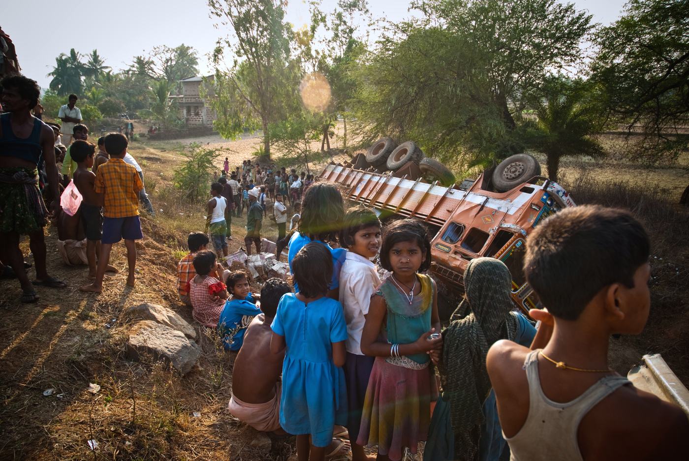 children and villagers gathered on a slope, some looking at the camera, an overturned truck lying behind them