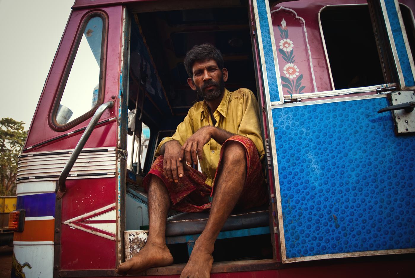a barefoot man in a yellow shirt sits in the open door of a painted red and blue truck cab