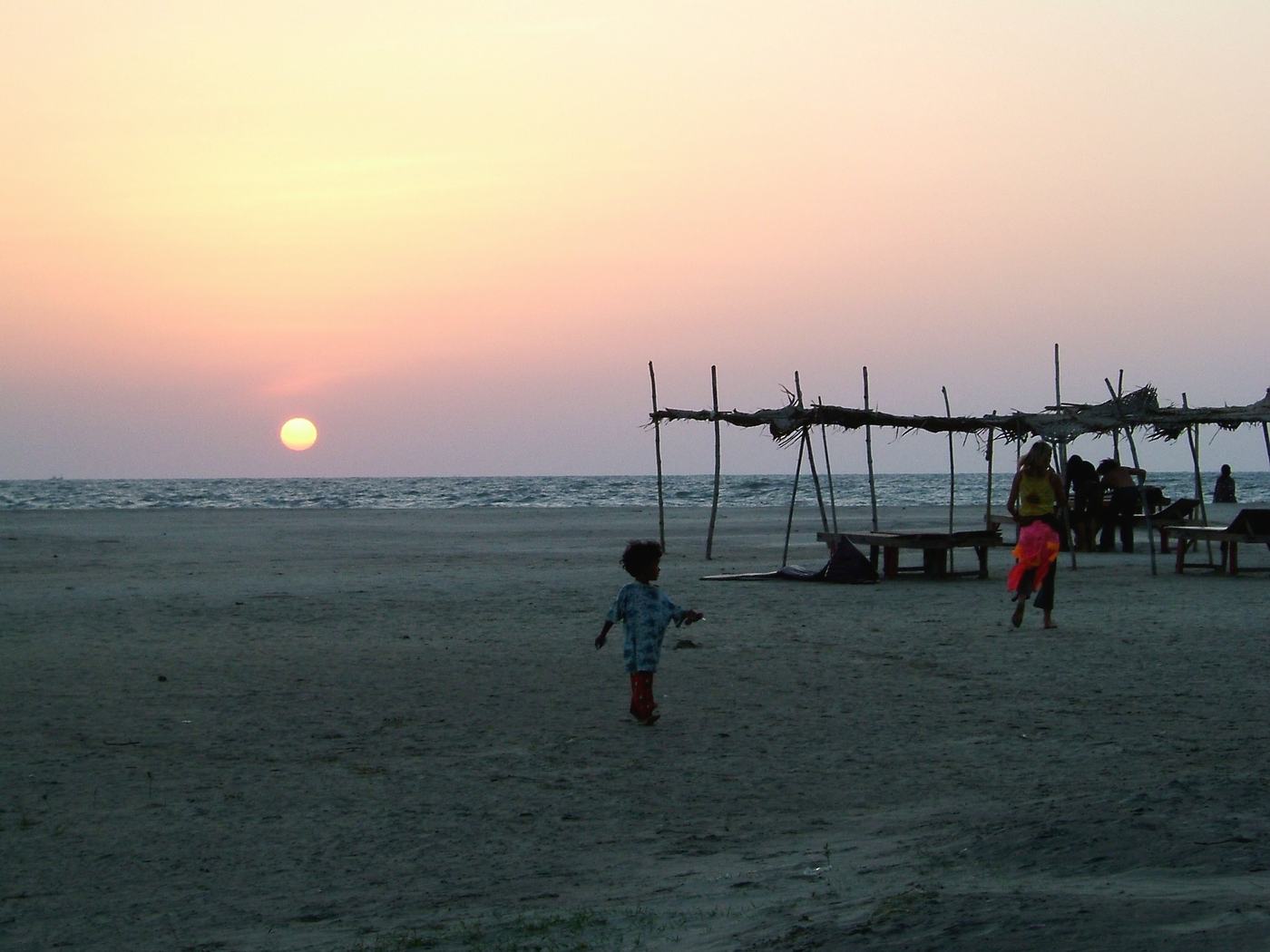a small child runs across a beach at sunset, the sun low over the sea, a frame of poles behind