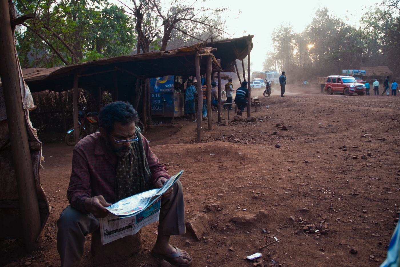 a man in glasses sits reading a newspaper beside a red dirt road lined with thatched stalls