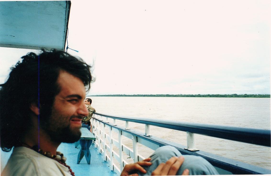 A smiling bearded man leans back at a boat railing with the wide Amazon behind him.