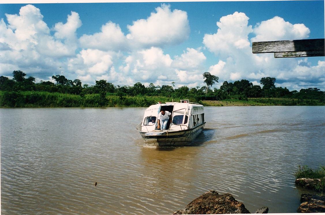 A small white passenger boat crosses the brown river outside Leticia under bright clouds.