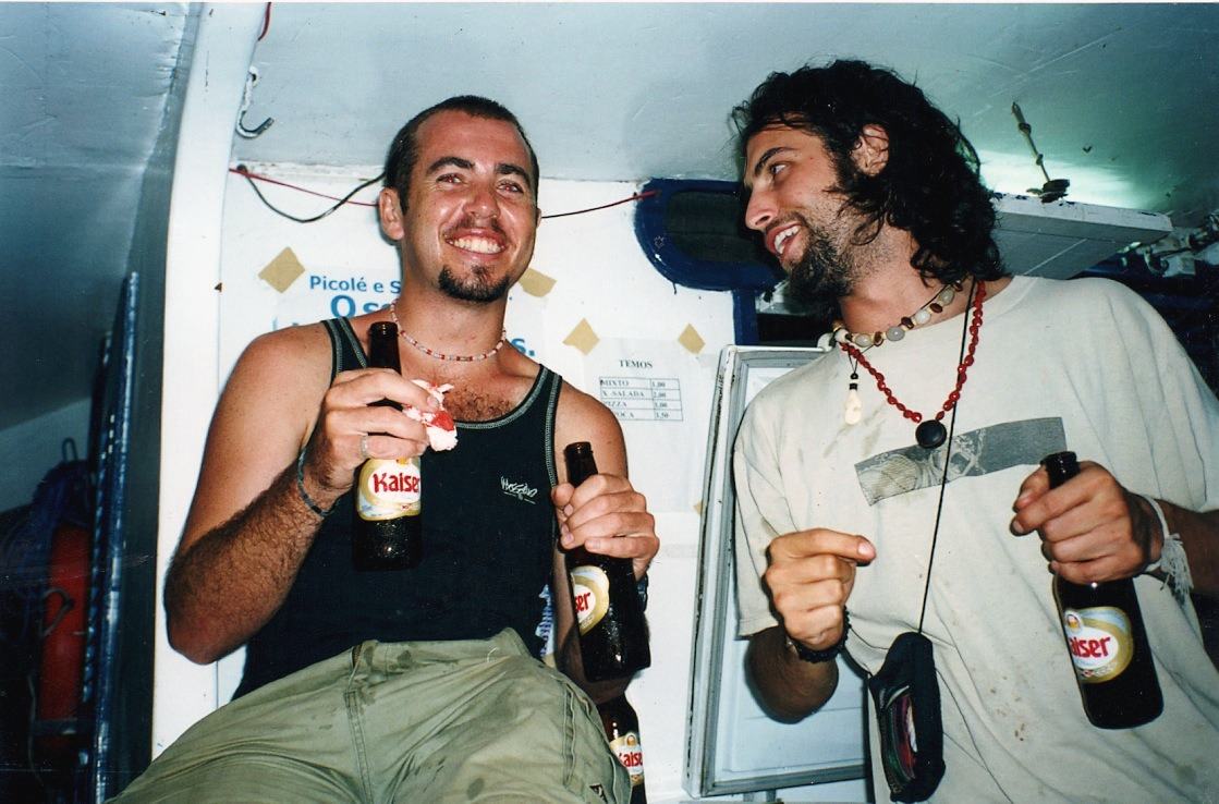 Two young men sit laughing in a boat cabin, each holding beer bottles.