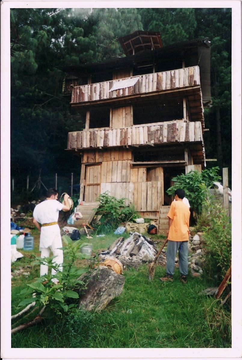 Two men stand in a green yard below a tall weathered wooden house with open upper floors.