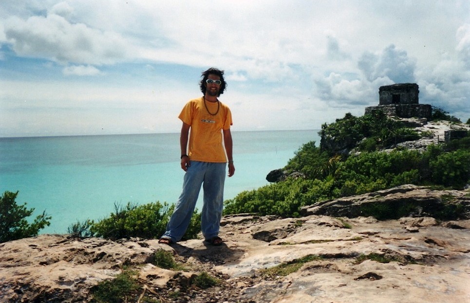 A young man in a yellow shirt and sunglasses stands on the rock above a turquoise sea at Tulum, a Mayan temple on the headland behind him