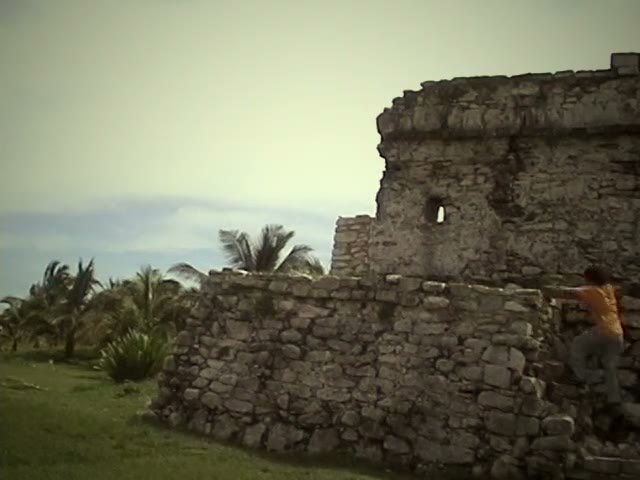 A still from video tape: a person in an orange shirt climbs the corner of a ruined grey stone building, palms and a pale sky behind.