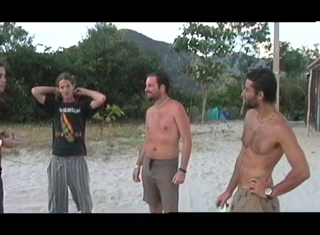Three young men on a beach, shirts off, a green headland behind them, a frame from the tape.