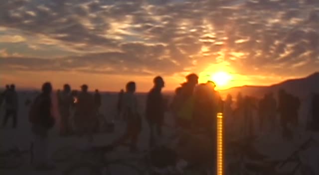 A crowd in silhouette on the desert floor at sunset, a low sun burning through a bank of cloud.