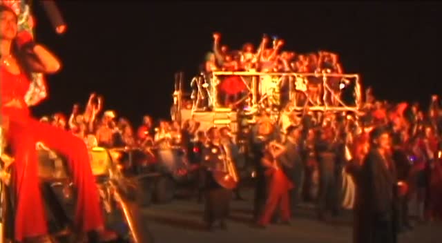 A night crowd lit red and orange, arms raised, a lit scaffold stage above them.