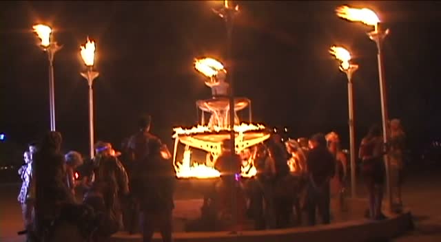 Torches on tall poles burning in the night, a crowd gathered round a lit structure.