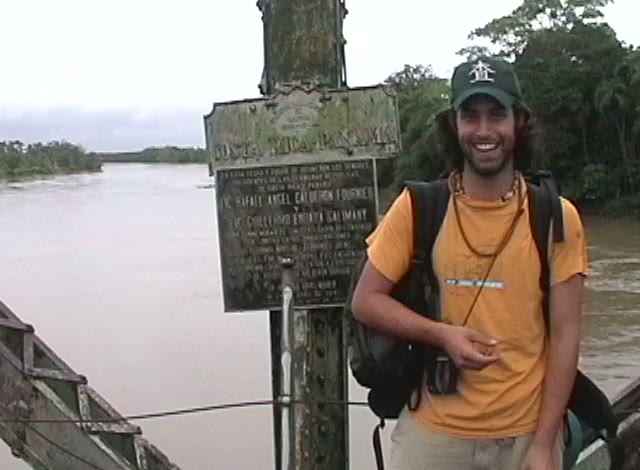 A young man in a cap and orange shirt smiling beside a sign at a wide brown river.