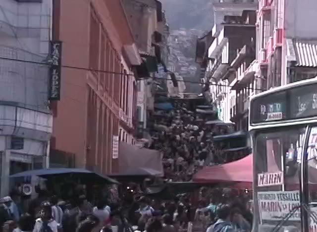 A crowded street market in a city, awnings and shop signs, people filling the road between buildings.