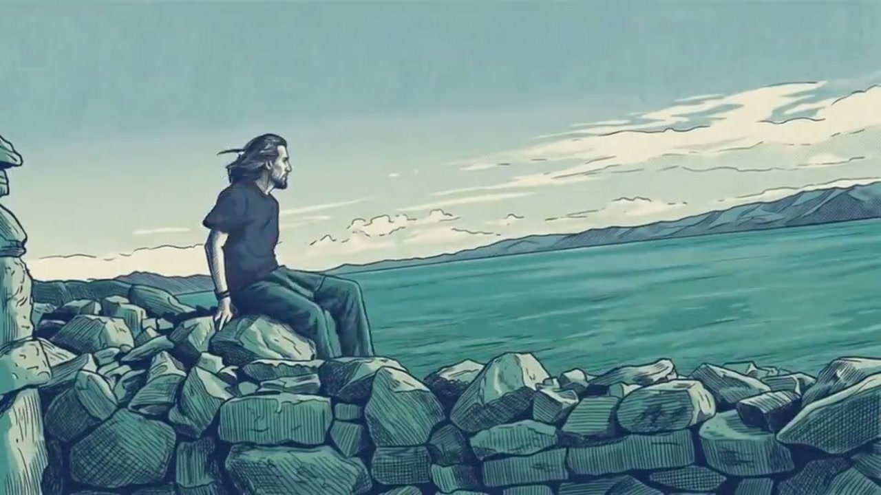 A long haired man in a black T shirt sits on a rough stone wall looking out over a wide green lake to a mountain range under clouds.