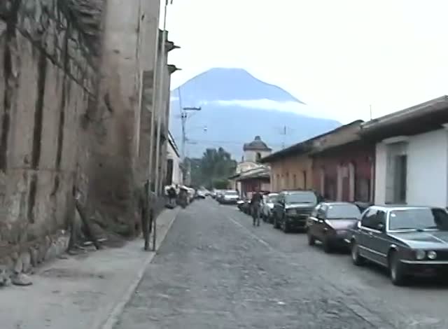 A cobbled colonial street of low houses, parked cars, a volcano rising into the clouds at its end.