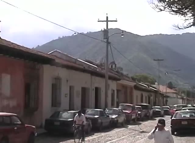A cobbled street of low houses under a green mountain, parked cars and a man on a bicycle, a frame from the tape.