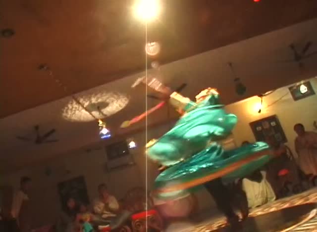 A dancer in green spinning under a bare light bulb in a crowded room at night.