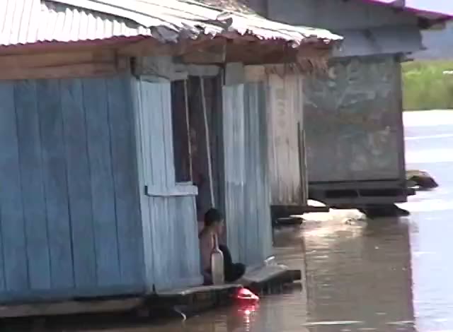 Blue plank houses on stilts at the river's edge, a woman sitting in a doorway above the water, a frame from the tape.
