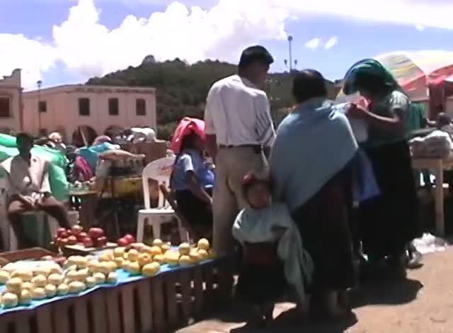 A market square, piles of fruit on a blue cloth, women in shawls and a child in the crowd, a church behind, a frame from the tape.