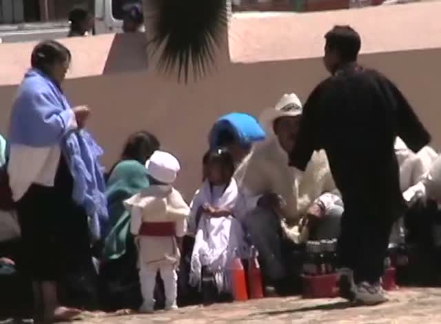A crowd at a village fiesta, people in blue shawls and white, children in the foreground.
