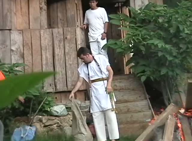 A man in white by a wooden house and steps, another standing above on the porch, green leaves in front.
